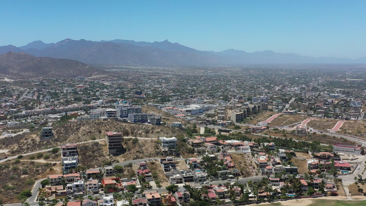 Opening aerial frame over San Jose del Cabo showing hillside homes and mid-rise buildings in front of a broad city grid and blue mountain horizon, with no visible people.