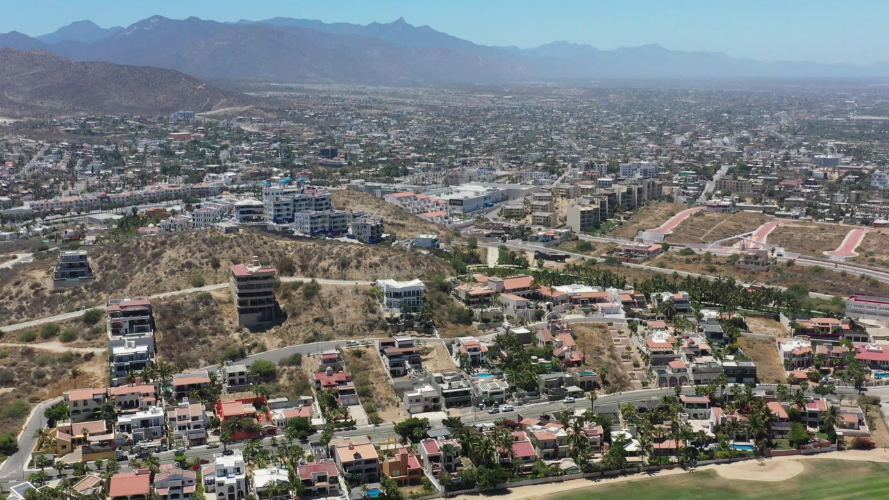 Wide opening view over San Jose del Cabo with hillside residences, dry slopes, a green fairway edge, a broad city grid, hazy mountains, and no visible people.