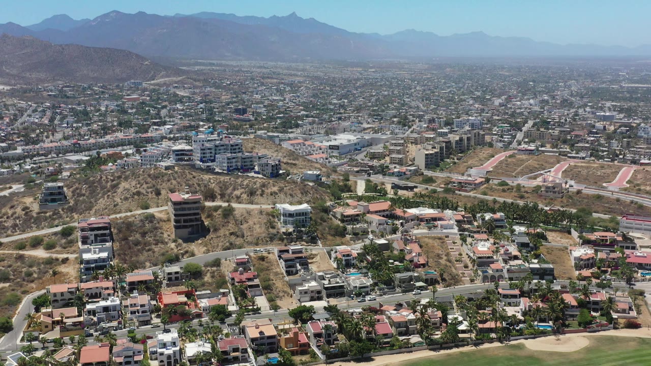 San Jose del Cabo hillside neighborhood seen from above, with modern houses, tan undeveloped slopes, curving streets, distant urban blocks, mountains, and no visible people.
