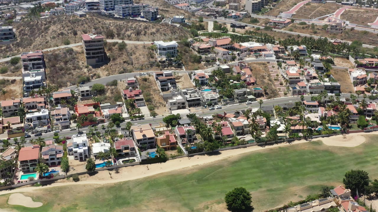 Aerial view in San Jose del Cabo with a broad fairway below, palm-lined residences along the road, small vehicles, pools, and hillside apartments in the distance, with no visible people.