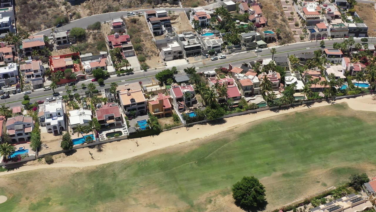 High overhead view in San Jose del Cabo with houses and pools along a road, a narrow sand strip, and a broad green open area in the foreground.
