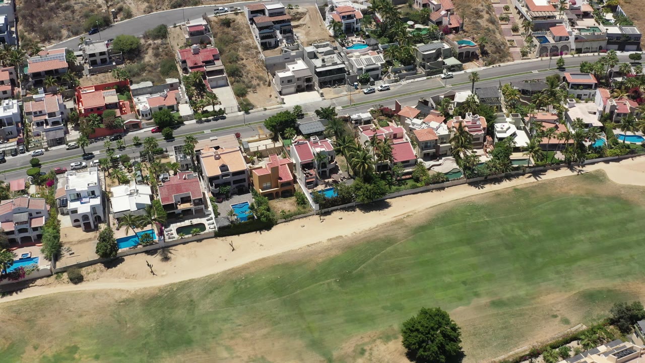 Aerial frame over San Jose del Cabo showing red and white roofed homes, parked and moving cars, palms, sand, and green ground below.