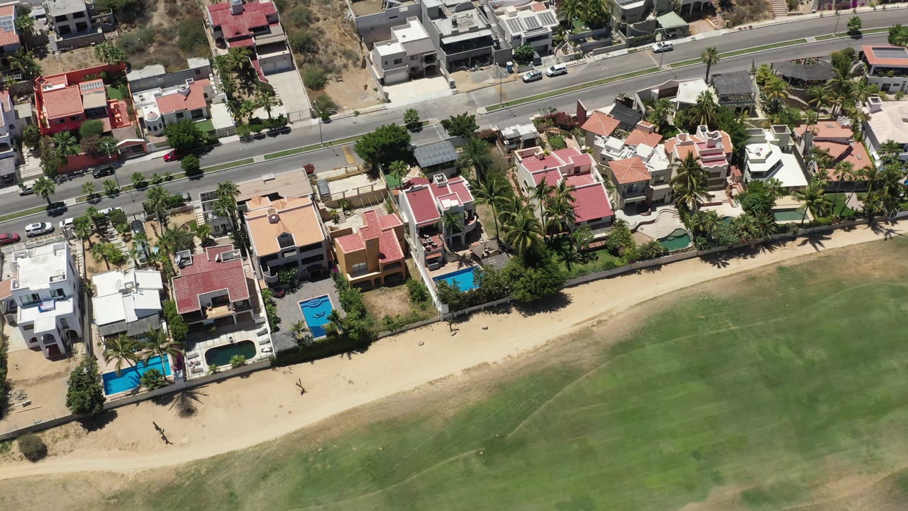 Drone-height view in San Jose Del Cabo with beachside houses, small blue pools, palm trees, a paved road with cars, sandy shore, green water, and no visible people.