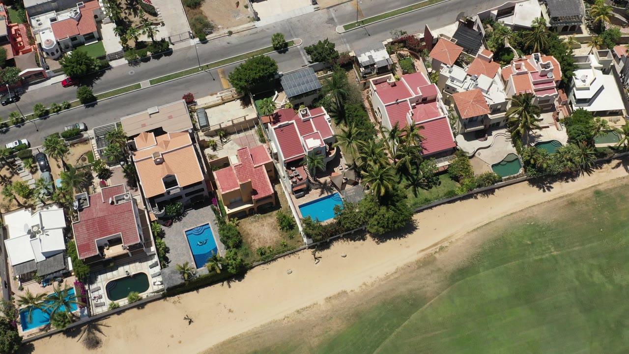 Top-down aerial view of beachside houses, road, pools, sand, and shallow water.