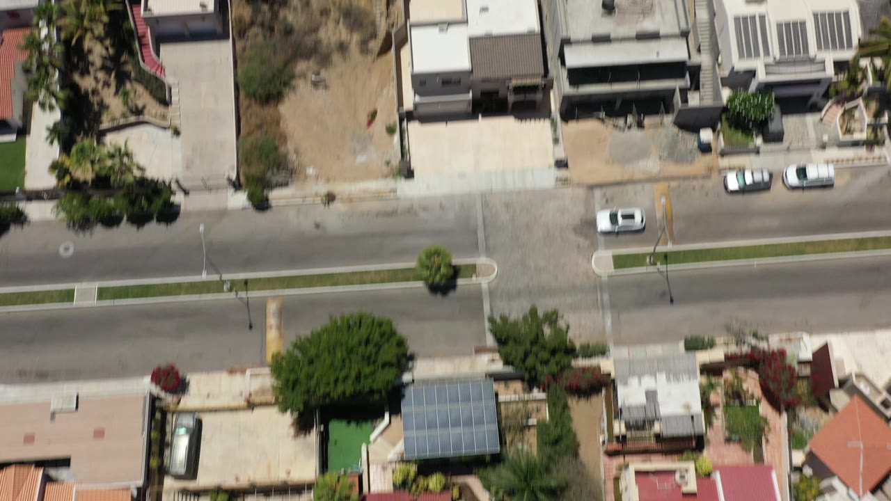 Aerial frame over San Jose del Cabo homes and a straight road, with several cars near driveways, dry undeveloped lots, and no visible people.