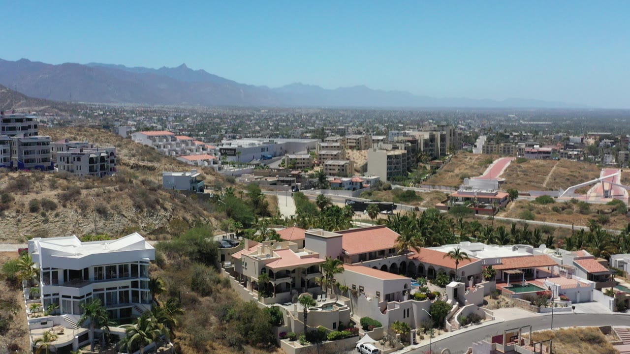 Opening frame over San Jose del Cabo hillside homes with red roofs, dry brush, palms, city buildings, blue sky, distant mountains, and no visible people.