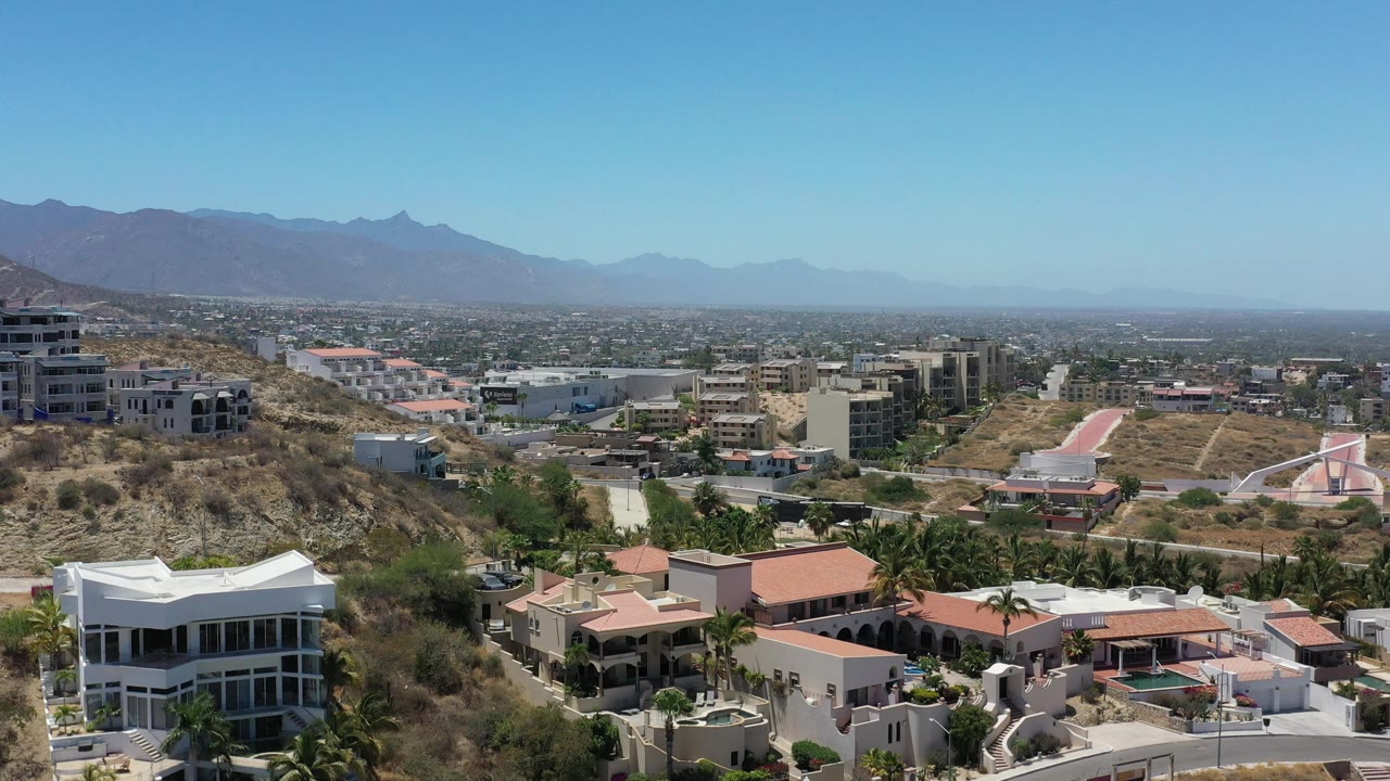 Residential rooftops and palms below a wide blue sky and mountain range.