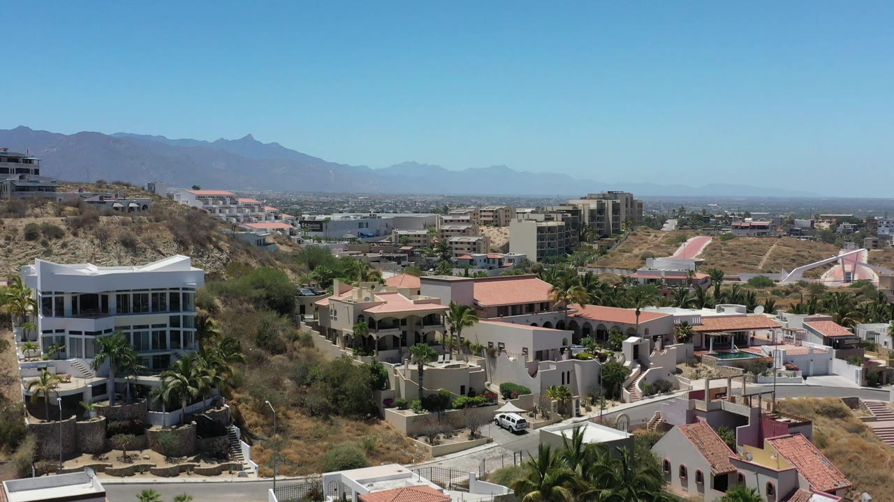 San Jose Del Cabo residential hillside seen from above, showing a white modern house at left, red roof villas below, palms, roads, and mountains in the distance, with no visible people.
