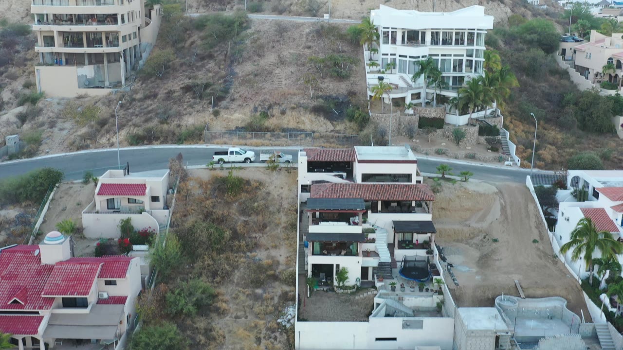 San Jose Del Cabo hillside view with a rounded beige apartment building, white multi-story residences, dry scrub slopes, a paved road, and no visible people.