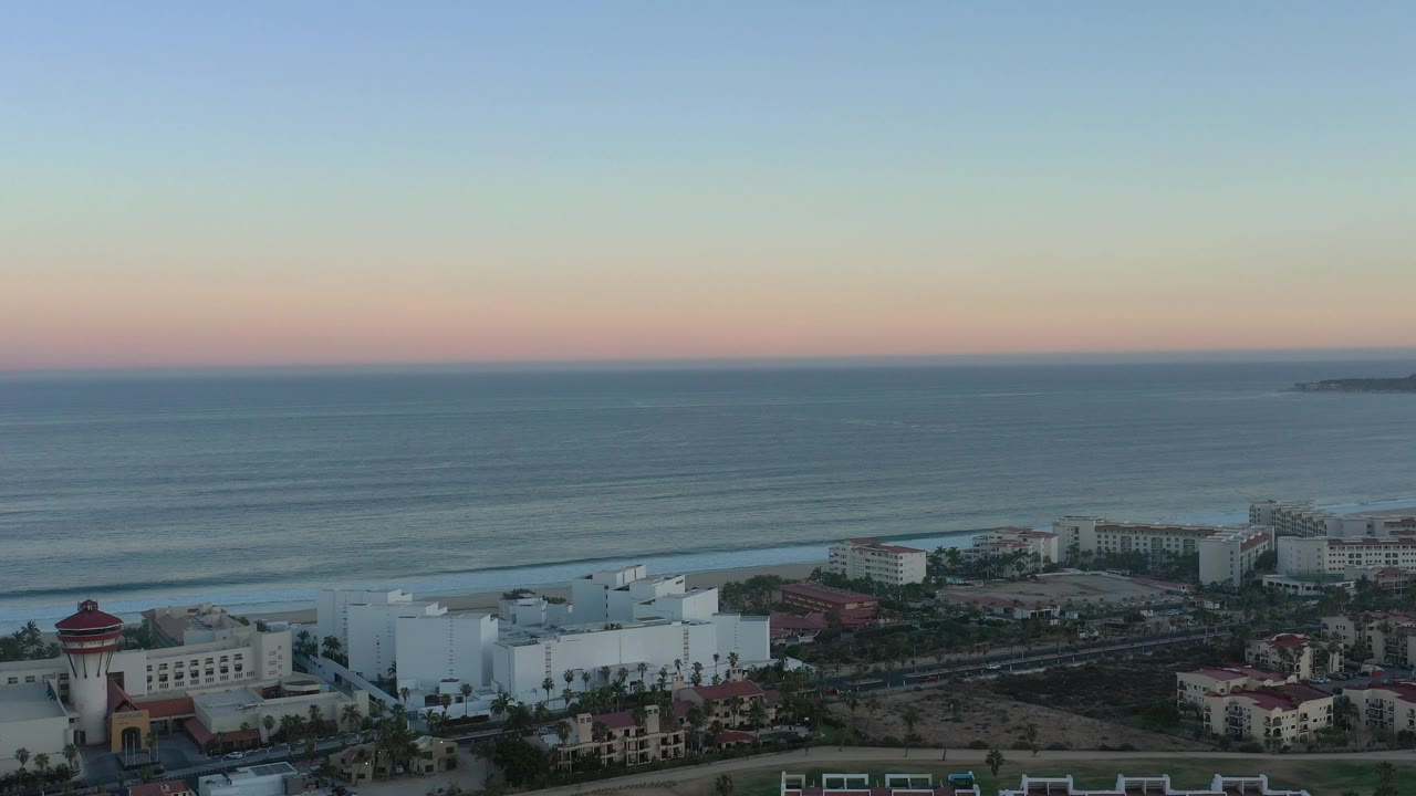 Aerial view of coastal buildings, beach, ocean waves, and sunset sky.