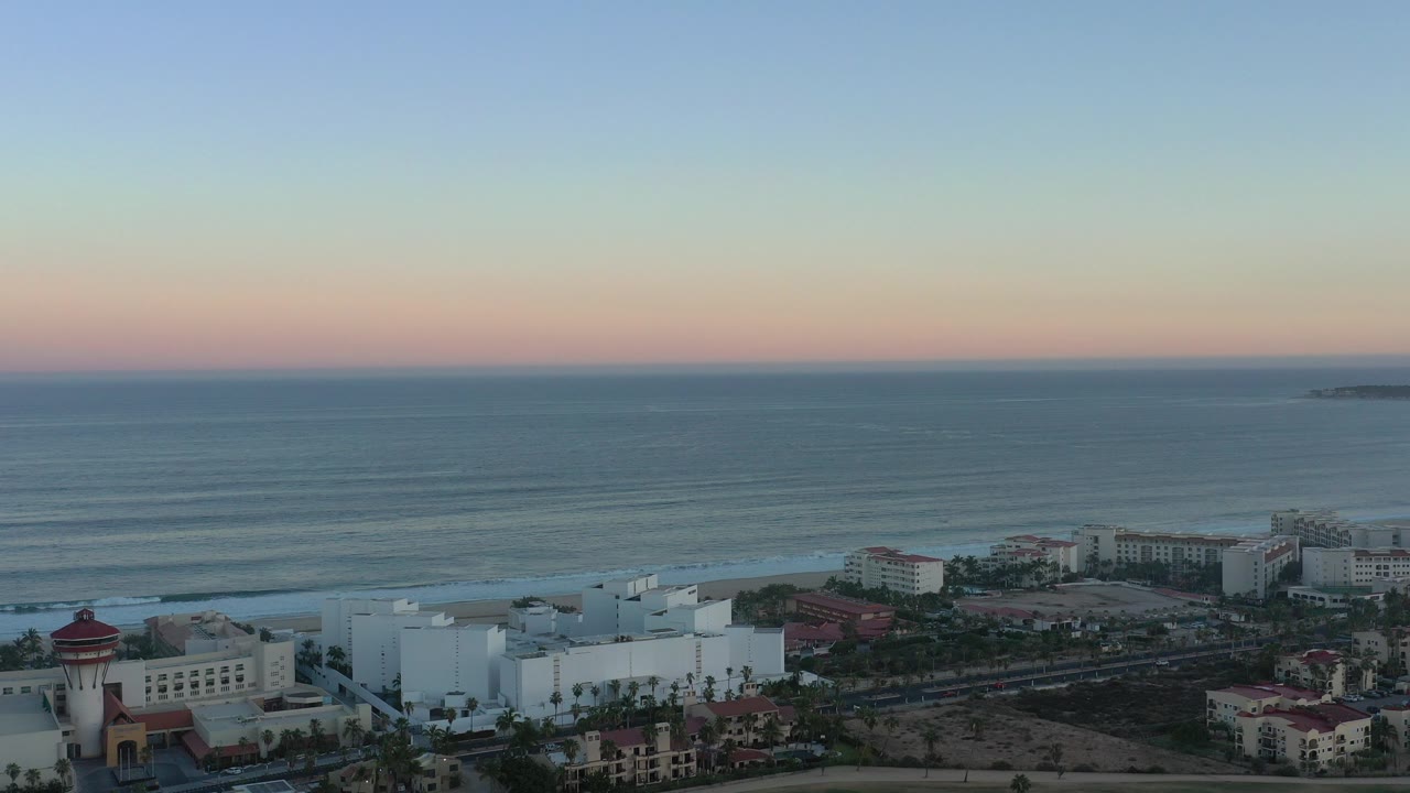 Wide aerial view over San Jose del Cabo hotels beside the ocean, with a pastel horizon, low surf lines, and no visible people.