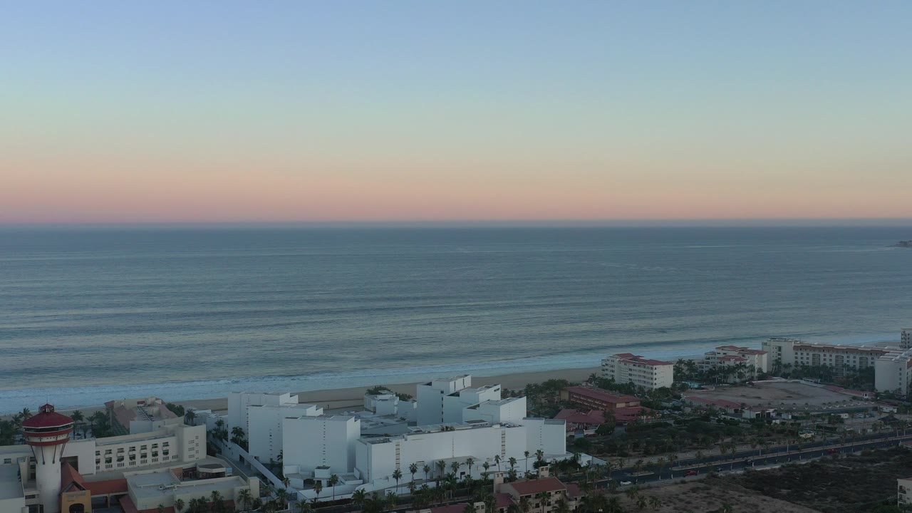 Wide aerial view in San Jose del Cabo, Mexico, showing a pastel dawn horizon over the sea, white coastal buildings at the lower edge, and no visible people.