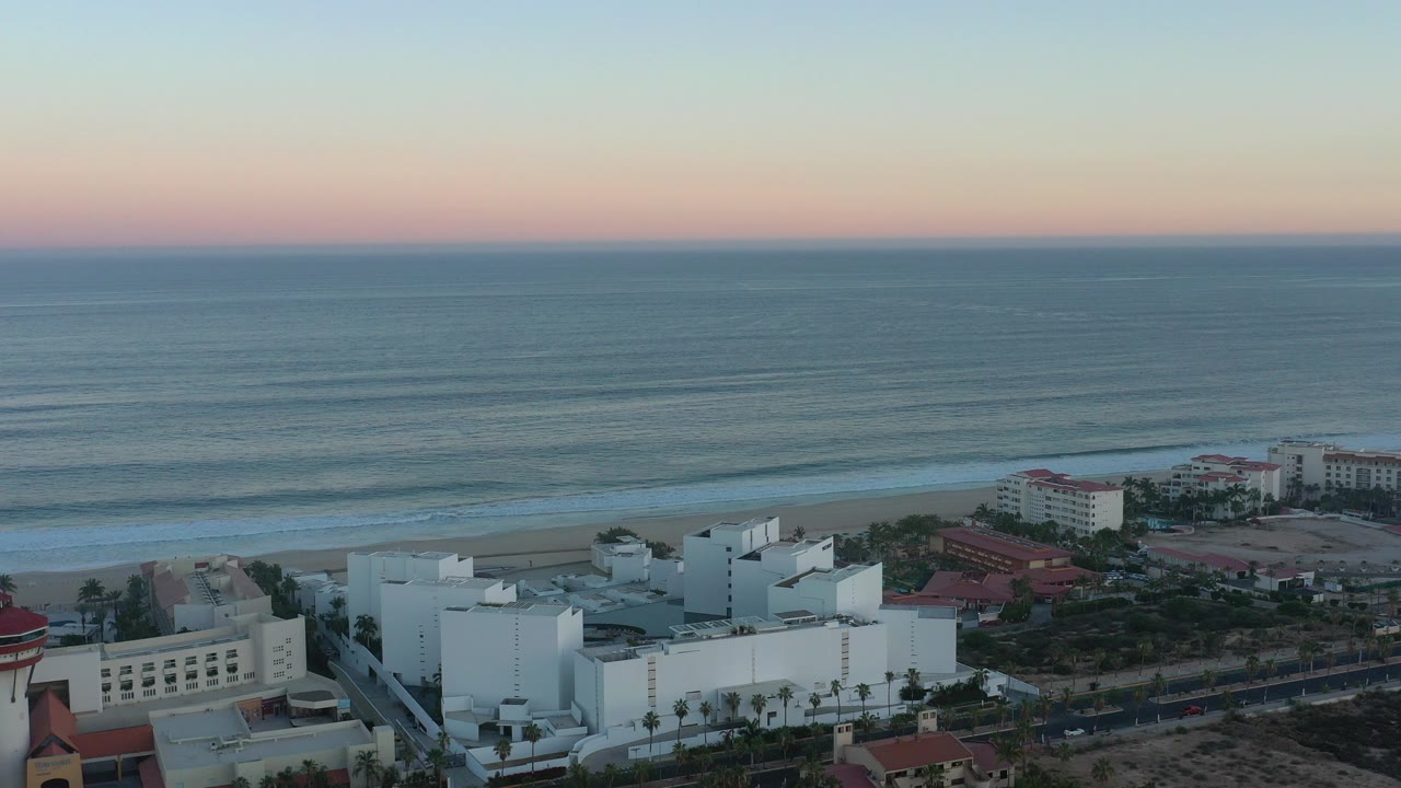 Opening aerial view in San Jose del Cabo with white coastal buildings below, a narrow sandy beach, lined ocean waves, a pastel horizon, and no visible people.