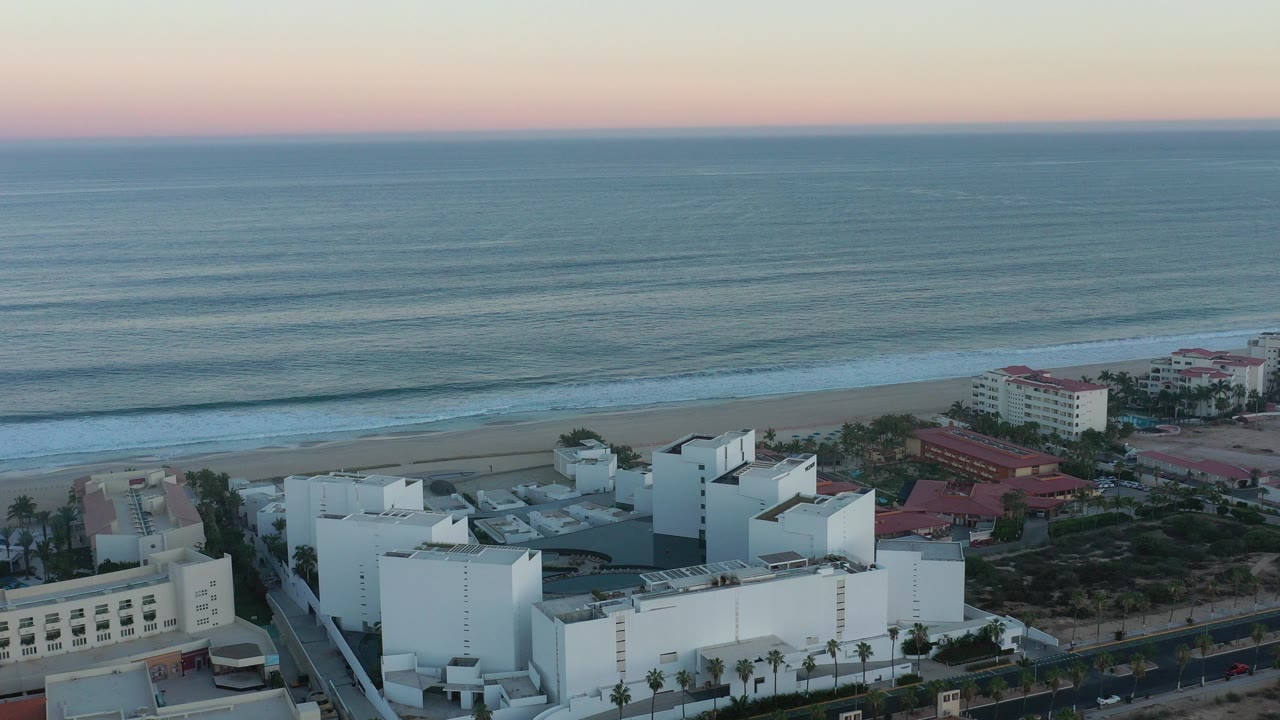 Opening elevated view of San Jose Del Cabo resort buildings, sandy beach, layered ocean waves, pastel sunset sky, and no visible people.