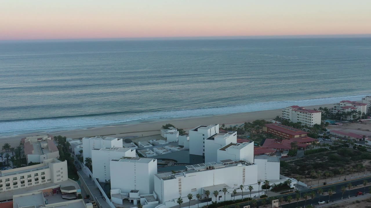 Elevated San Jose Del Cabo coastal view with white hotel roofs lower in frame, red resort structures to the right, ocean bands beyond, and no visible people.