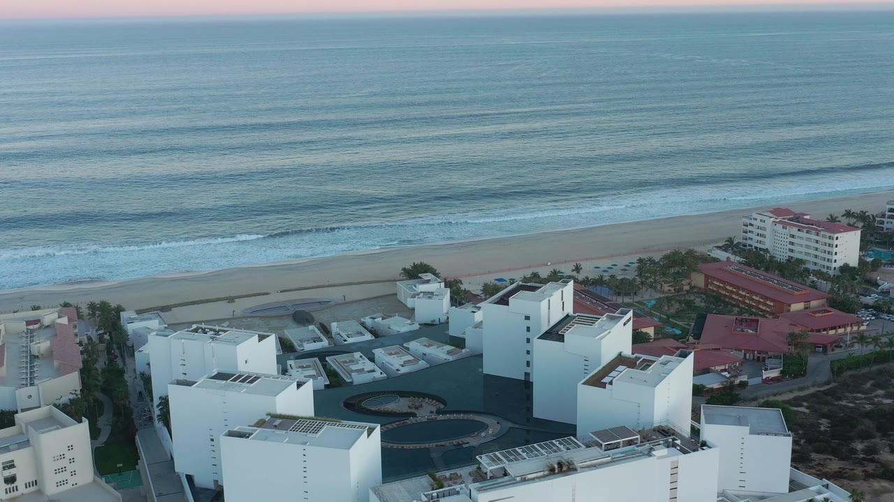 San Jose del Cabo aerial frame with flat-roofed white buildings, a circular dark paved feature, beachfront structures, repeating wave bands, and no visible people.