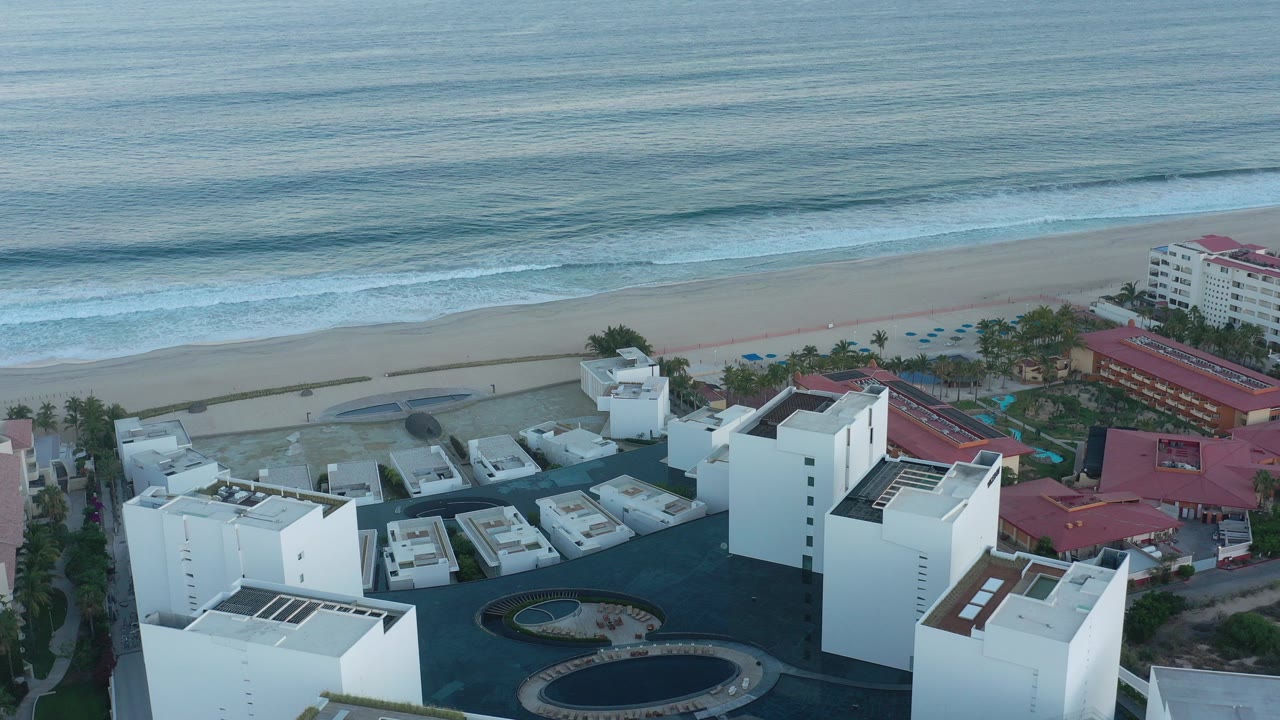 Aerial view of white waterfront buildings beside a sandy beach and surf.