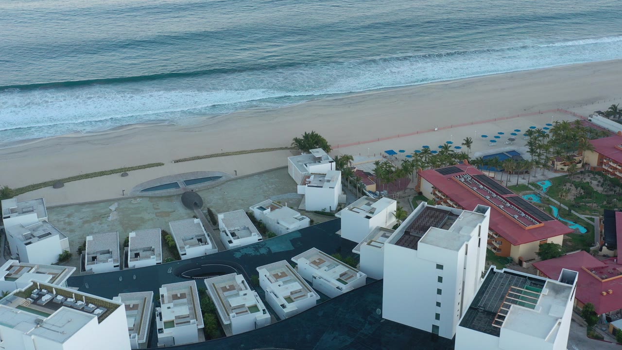 Aerial frame over San Jose Del Cabo resort buildings, with white rooftops in the foreground, a red-roofed hotel wing to the right, beach and blue surf beyond, and no visible people.