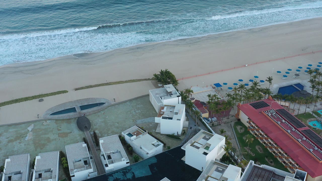 Aerial view of surf, sand, beachfront buildings, umbrellas, and palms.