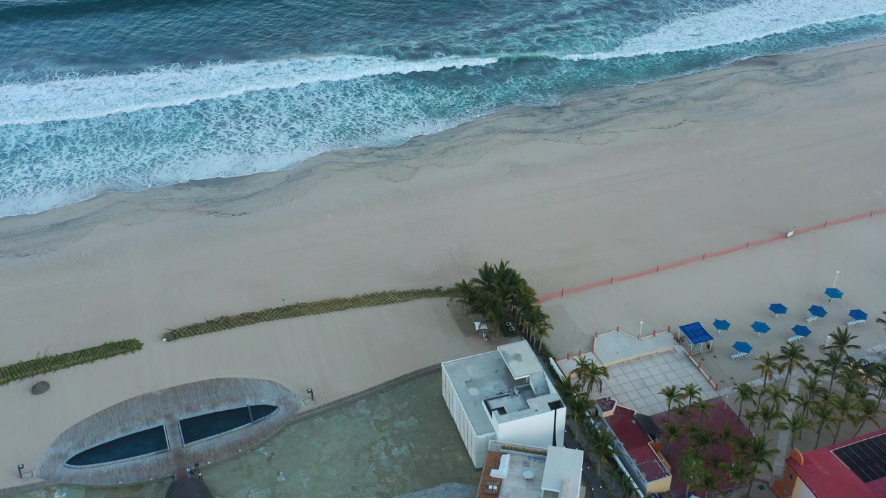 Overhead beach with surf, sand, palms, pool area, and blue loungers.