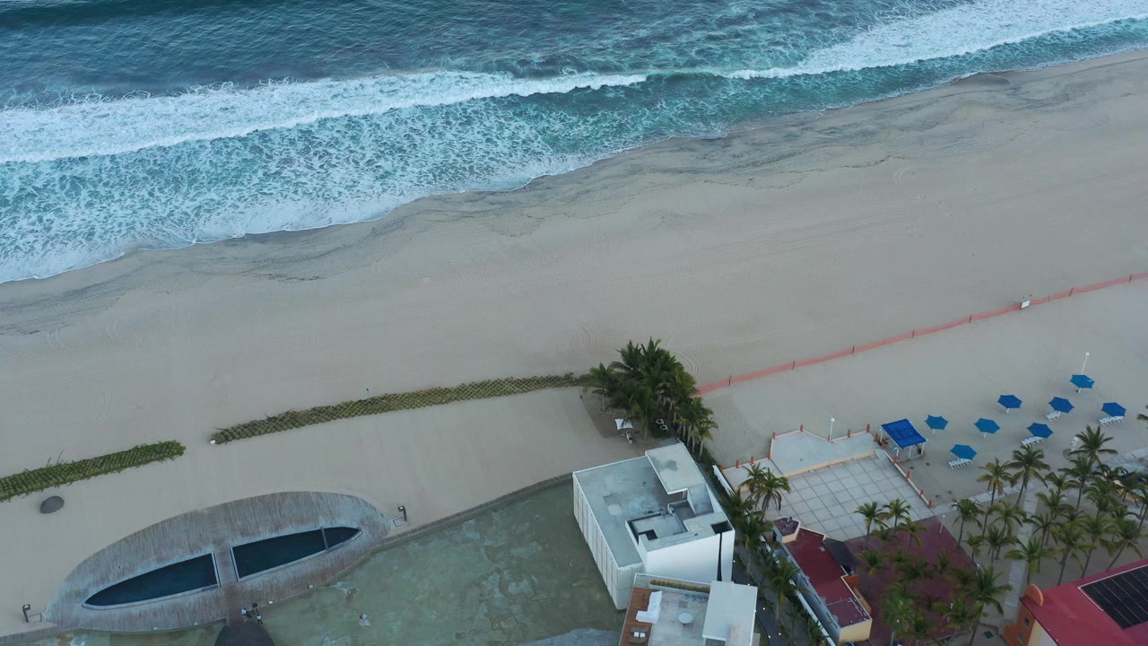 Aerial San Jose del Cabo shoreline view where foamy waves sit higher in frame and the beach, red line, palms, patio umbrellas, blue loungers, and pool remain visible with no visible people.