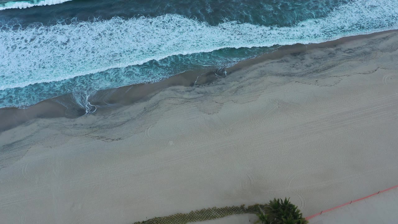 Aerial frame of white foam spreading along the upper beach in San Jose del Cabo, Mexico, with broad smooth sand below and no visible people.