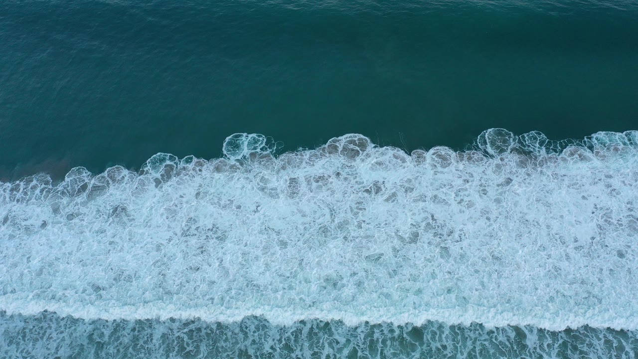 Top-down view of teal water and a broad white foam band breaking across San Jose del Cabo, Mexico, with no visible people.