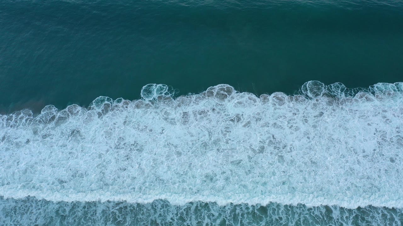 Aerial-looking view of white bubbly surf spreading across dark teal ocean water in San Jose del Cabo, Mexico, with no visible people.
