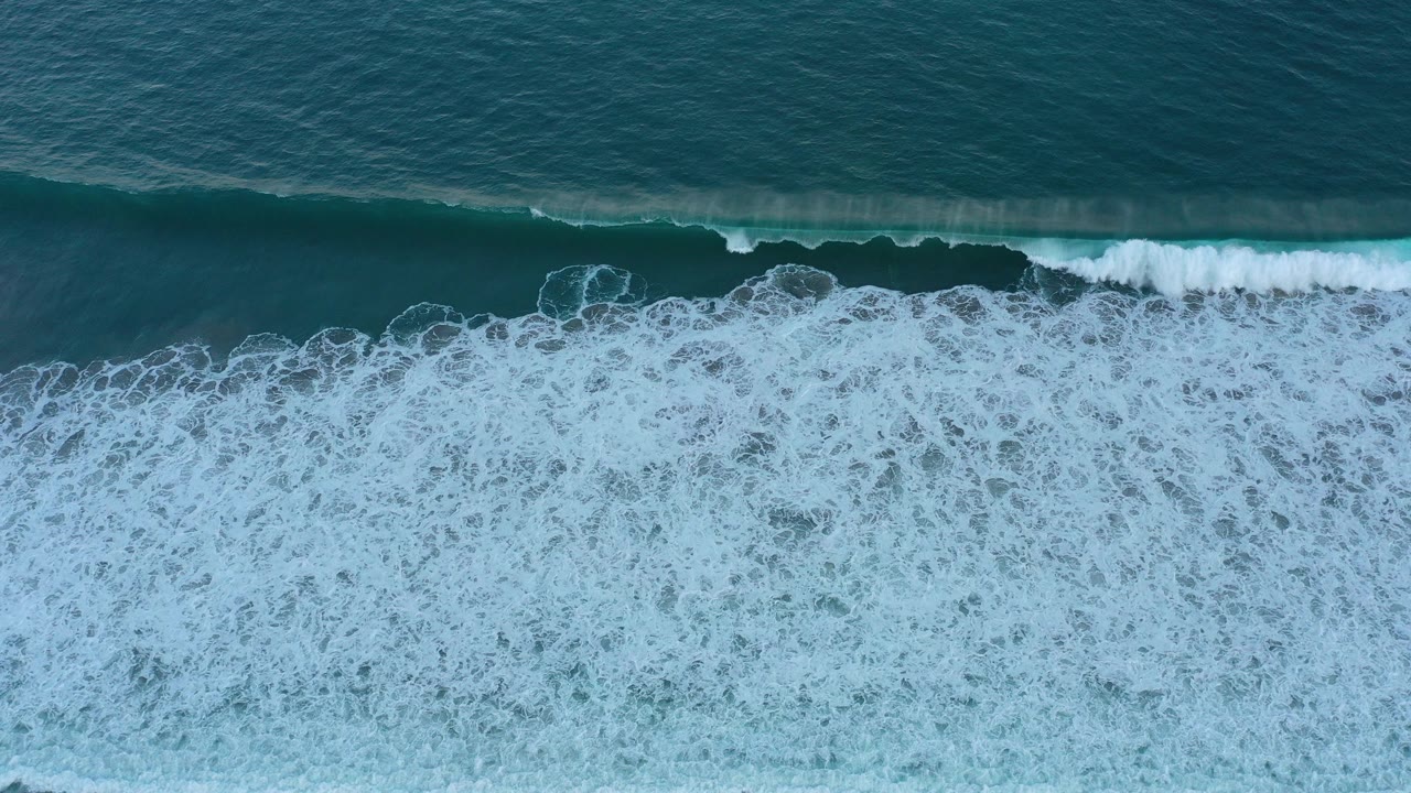 Overhead blue ocean wave breaking into white foam near sand.