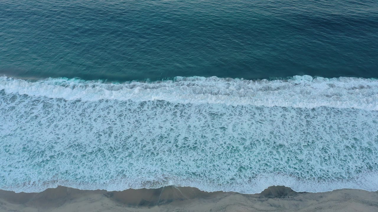 Overhead view of teal water and white surf meeting a sandy shoreline in San Jose Del Cabo, Mexico, with no visible people.
