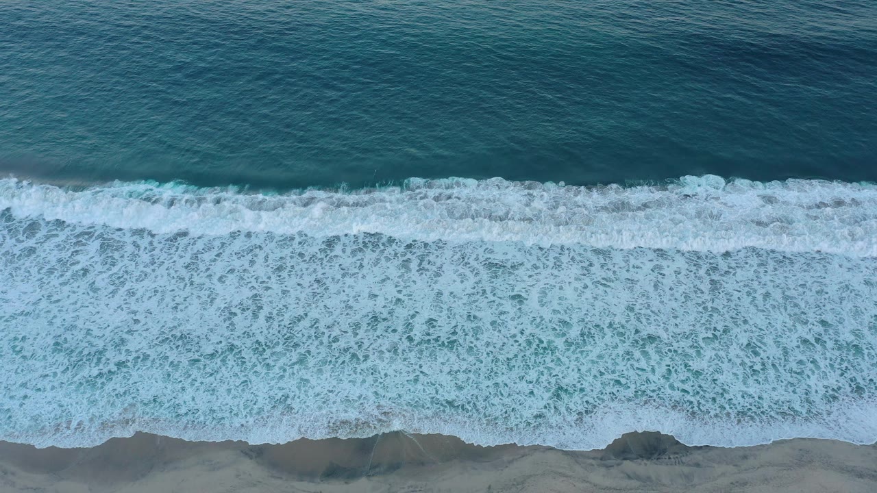 A later overhead frame shows the foam line shifted along the San Jose Del Cabo shoreline, with teal water above, wet sand below, and no visible people.