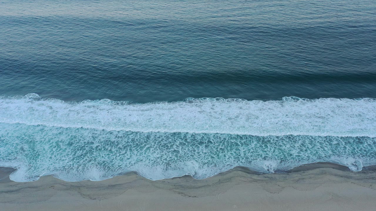 Overhead view of San Jose Del Cabo surf forming a white foam band across dark blue water and gray-beige sand, with no visible people.