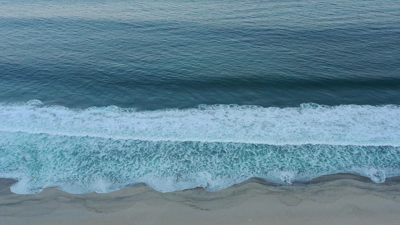 A broad white surf band spreads across the lower shoreline in San Jose Del Cabo, with darker open water above and no visible people.