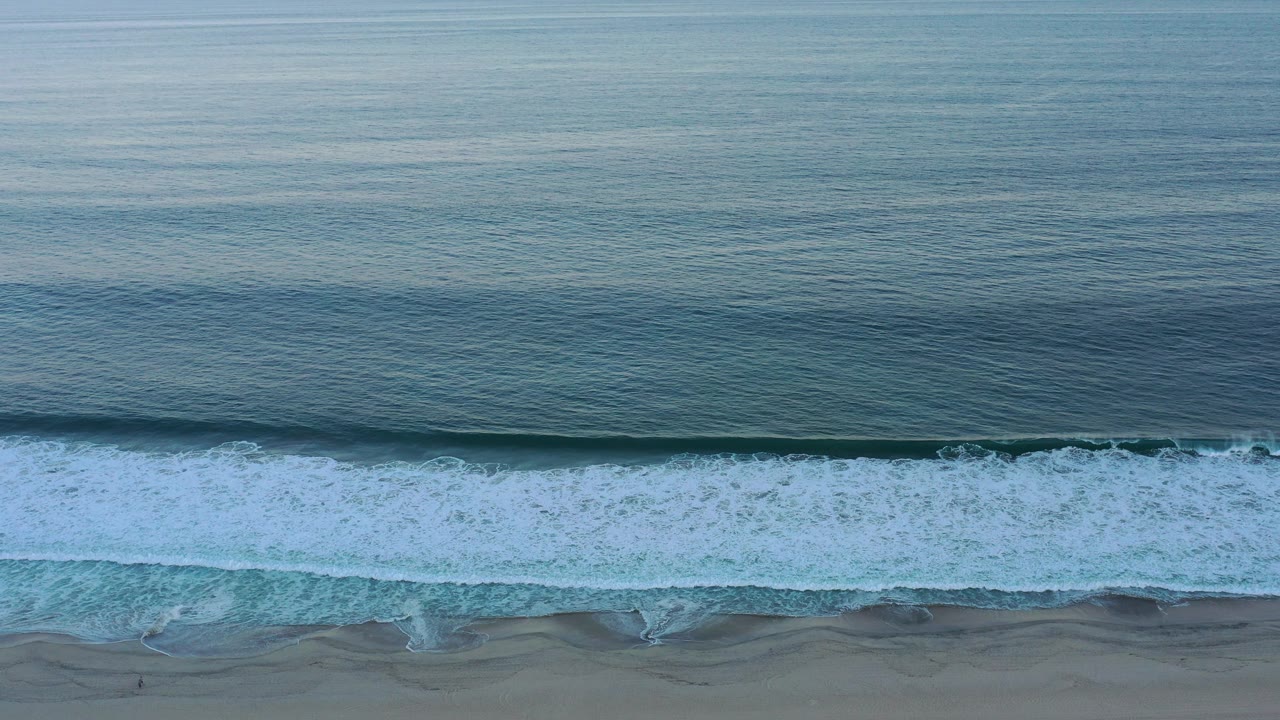 At 0:00, a wide elevated view in San Jose del Cabo shows blue ocean water, a long white wave line, wet sand at the bottom, and no visible people.