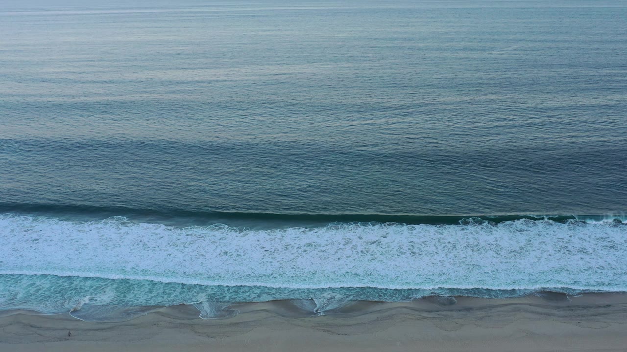 At 0:01, the breaking wave spreads farther across the lower frame while the calm blue ocean remains broad above it in San Jose del Cabo, with no visible people.