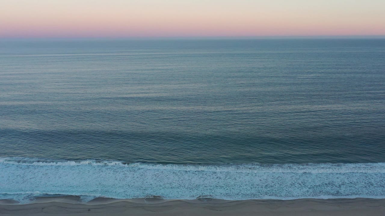 The San Jose del Cabo shoreline remains distant beneath a pink-blue sunset band, with small waves forming offshore and no visible people.