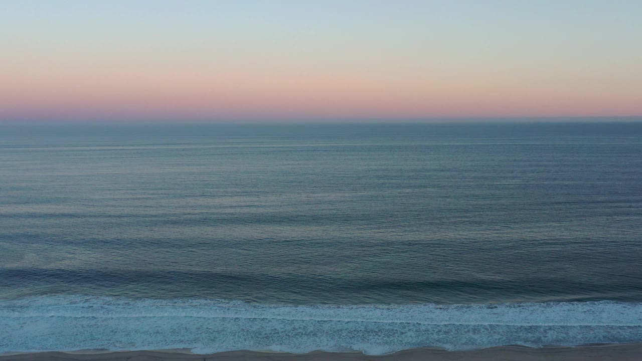San Jose del Cabo shoreline view with blue-green ocean, a pink band of sunset sky, white surf crossing the lower frame, and no visible people.