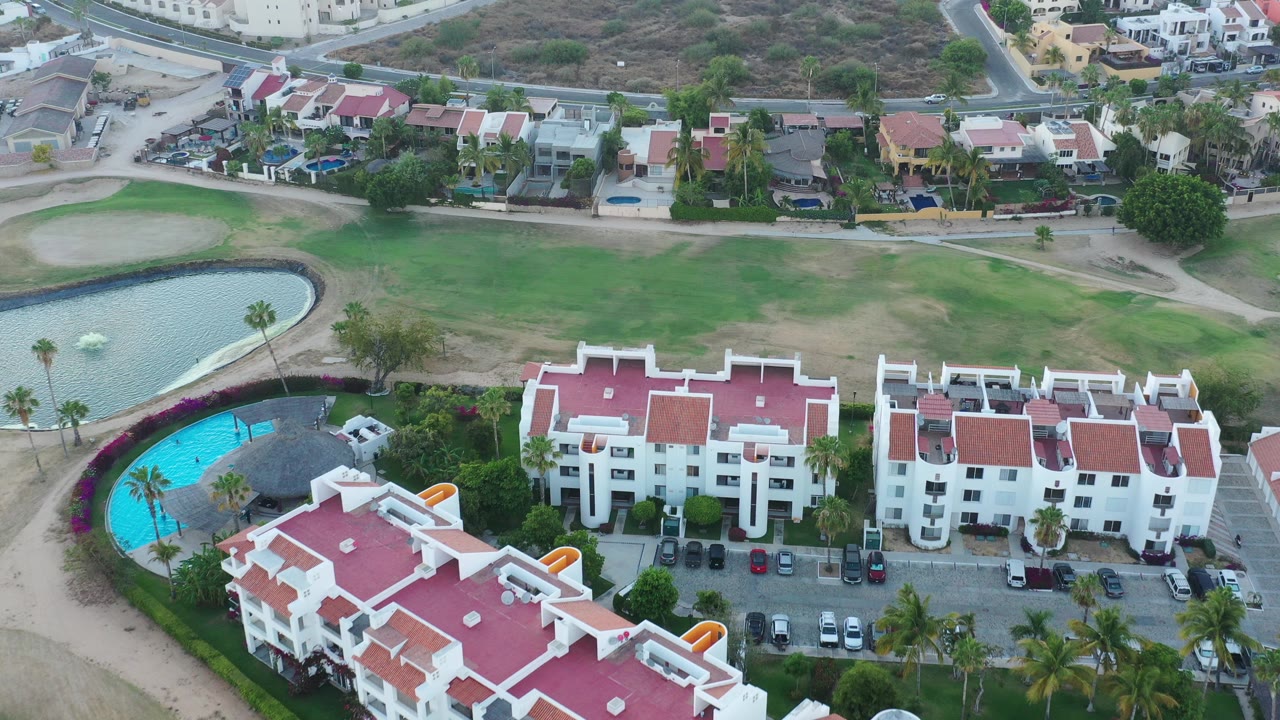 Aerial view of red-roofed white resort buildings beside a blue pool and lawns.