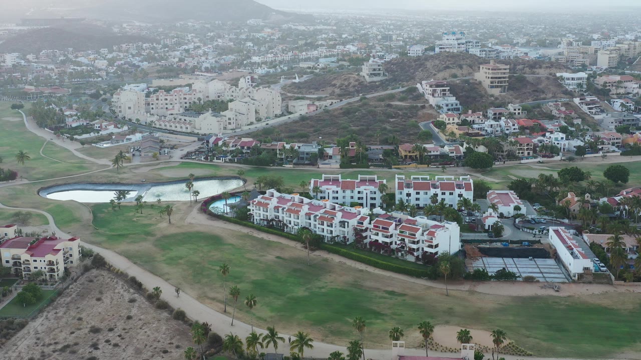 Aerial view over golf greens, ponds, red-roofed buildings, hillside homes, and hazy mountains.