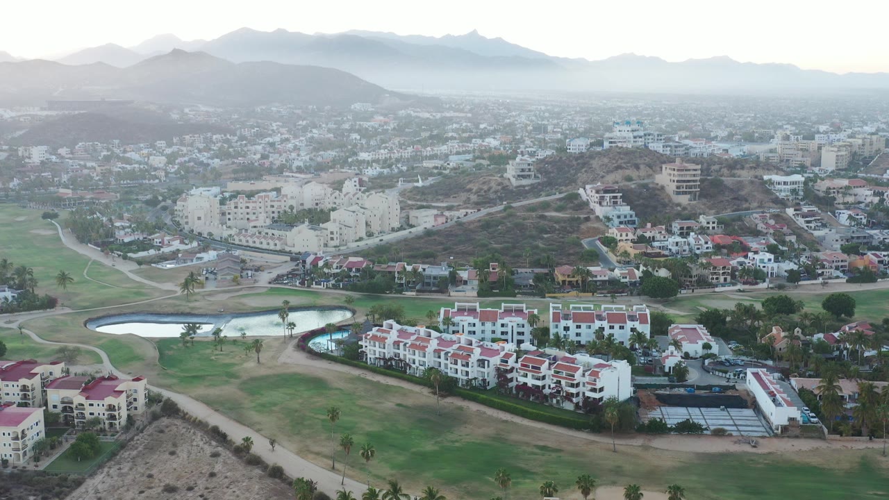 Aerial view of white homes, green grounds, ponds, roads, hills, and hazy mountains.