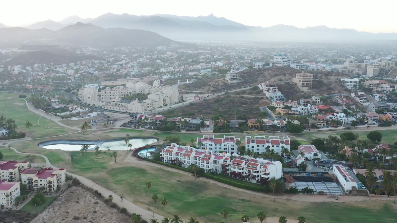 Second aerial frame in San Jose del Cabo, Mexico, with red-roofed residential buildings across the lower edge, ponds and green fairway-like grounds at left, hillside homes beyond, and no visible people.