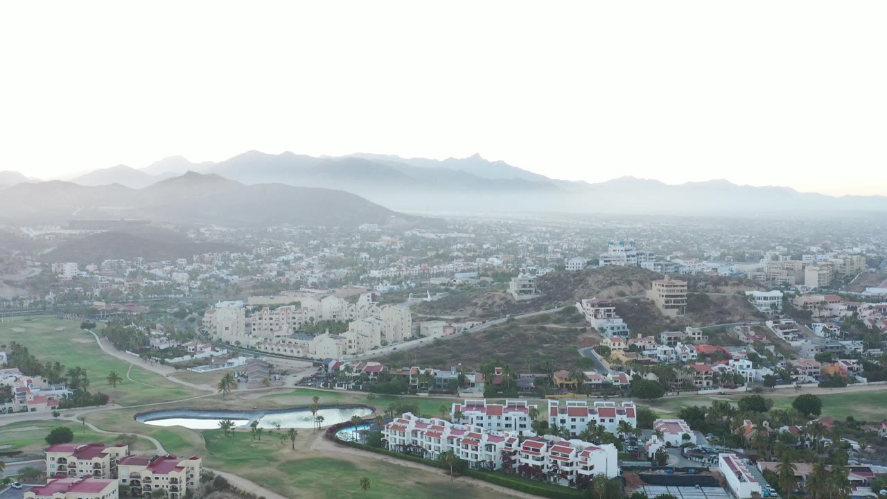 Aerial view over San Jose Del Cabo with green grounds, white buildings, small ponds, hazy mountains, pale sky, and no visible people at the opening frame.