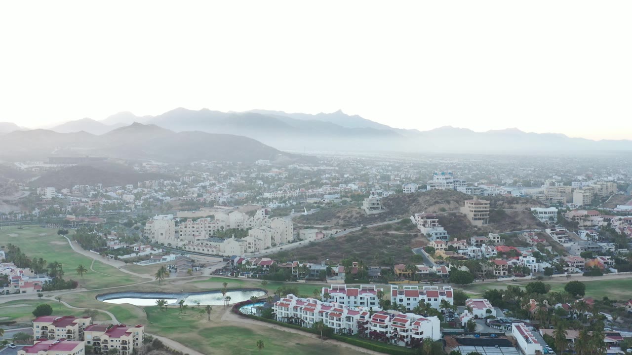San Jose Del Cabo appears in a broad aerial composition with red-roofed buildings in front, clustered town blocks beyond, layered mountains, fog, and no visible people.