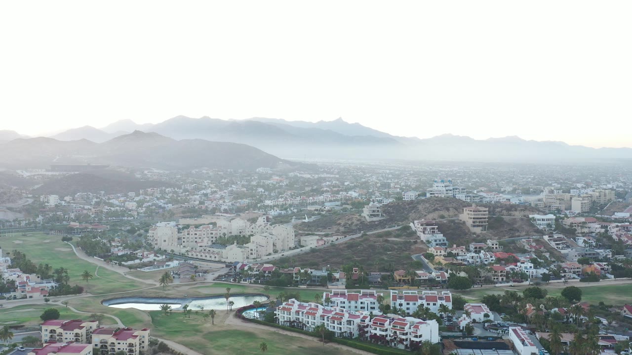 Elevated view across a hazy Mexican town with golf-like greens and mountains.