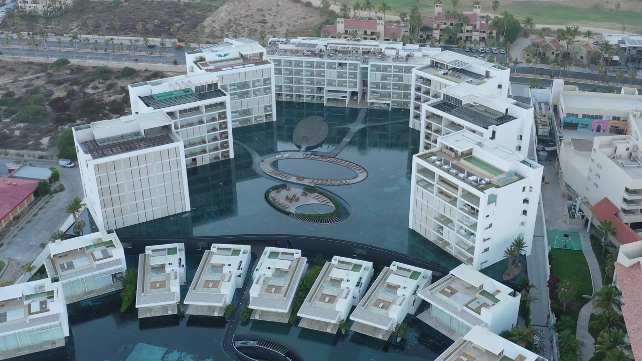 High aerial frame over a white building complex in San Jose del Cabo, Mexico, with dark water courtyards, foreground villa roofs, a background road, dry hillside, and no visible people.