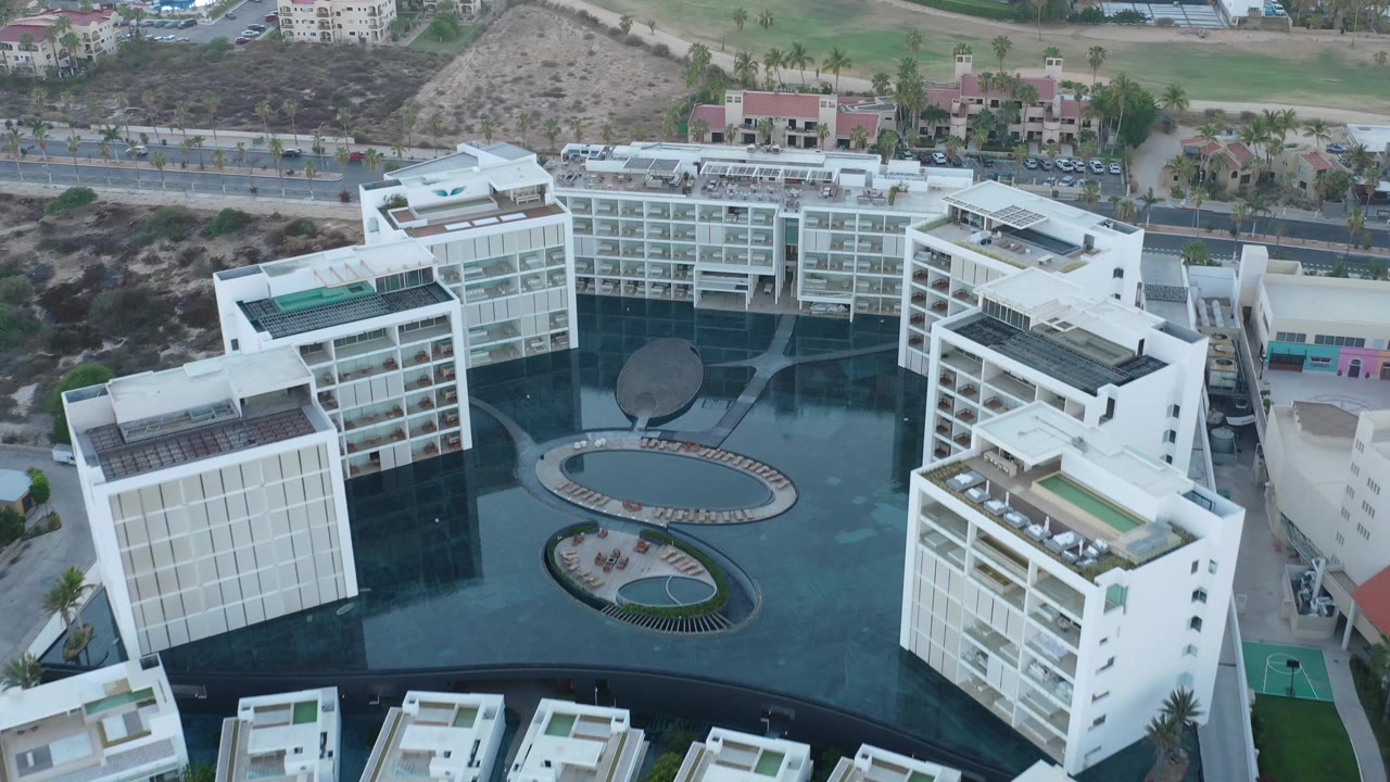 Aerial view in San Jose del Cabo showing white hotel wings around dark water features, with a road, scrub-covered hillside, green grounds, and no visible people.