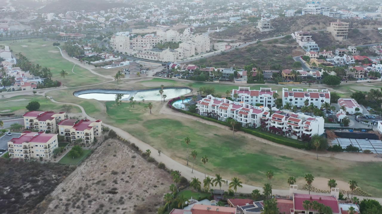 Aerial view in San Jose Del Cabo with two bright ponds near the center, white residential blocks with red roofs, open grass, hillside roads, distant buildings, and no visible people.