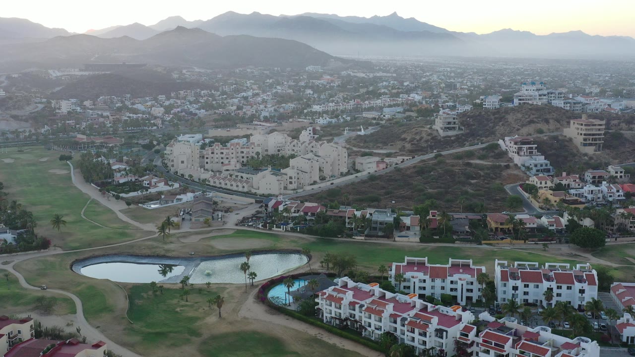 Aerial view in San Jose del Cabo, Mexico, with red-roofed white buildings beside golf-course ponds and layered mountains in the background, no visible people.