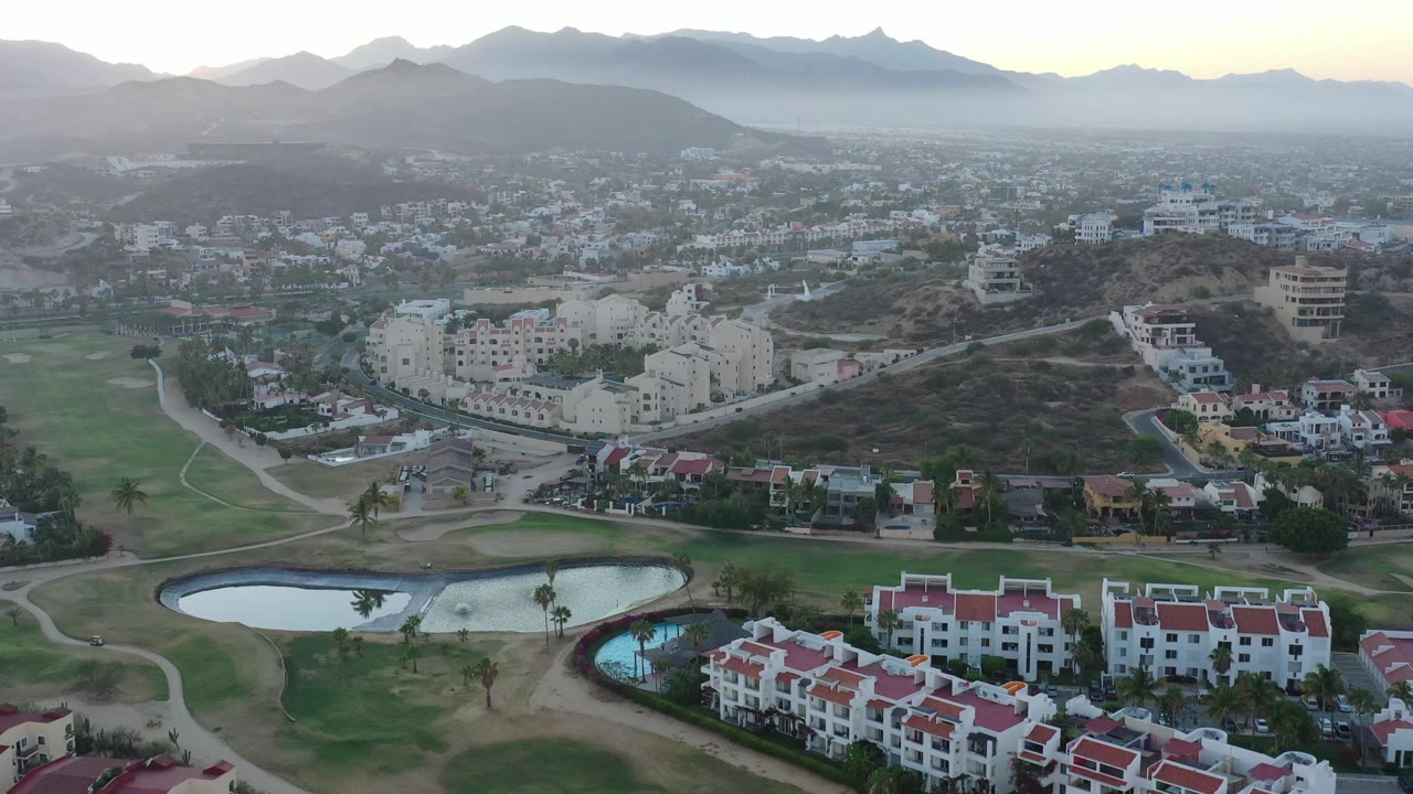 San Jose del Cabo aerial frame showing foreground ponds and white buildings with red roofs, a spread of city structures beyond, and hazy mountains under soft sky light, no visible people.