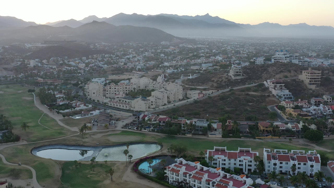 Wide aerial over a resort-like green area, housing, and mountains at sunrise or sunset.